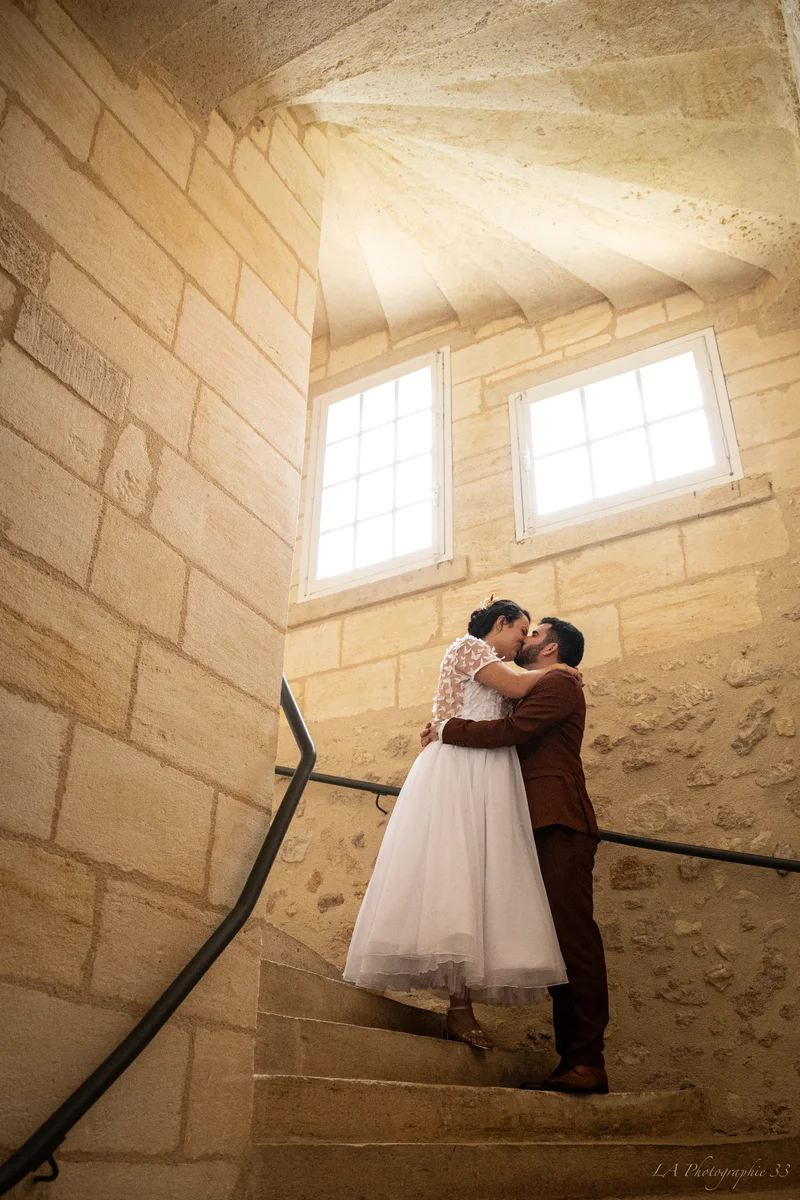 Mariés s'embrassant dans un escalier en pierre dorée, la mariée en robe blanche à appliqués papillons et le marié en costume bordeaux